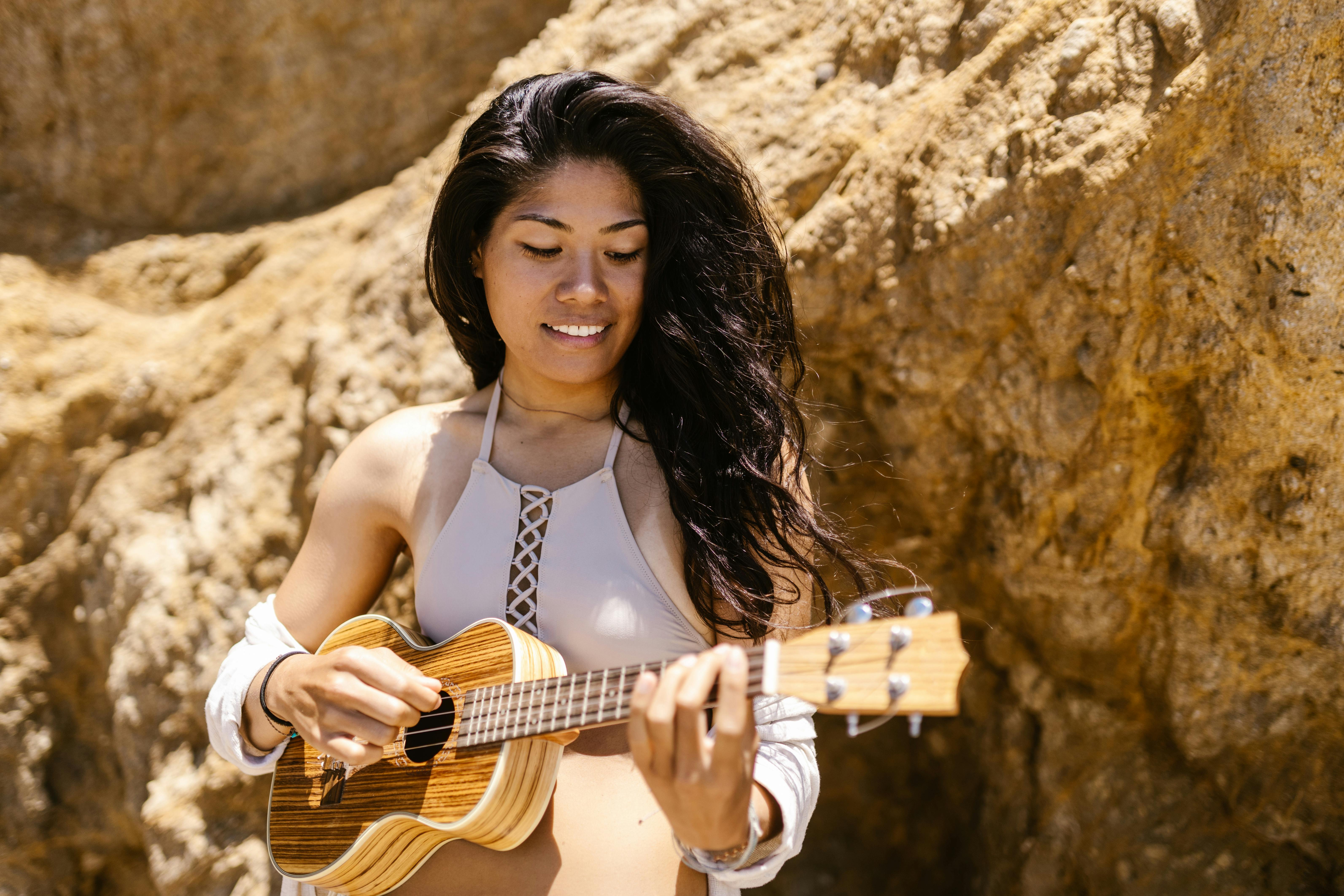 Smiling woman enjoying playing ukulele by a rock wall on a sunny day.