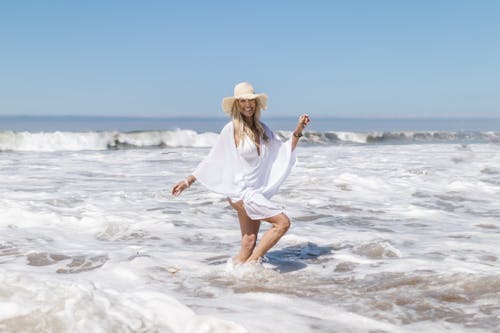 Free A Woman in White Bikini Wearing a Sun Hat Walking Through the Waves on the Beach Stock Photo