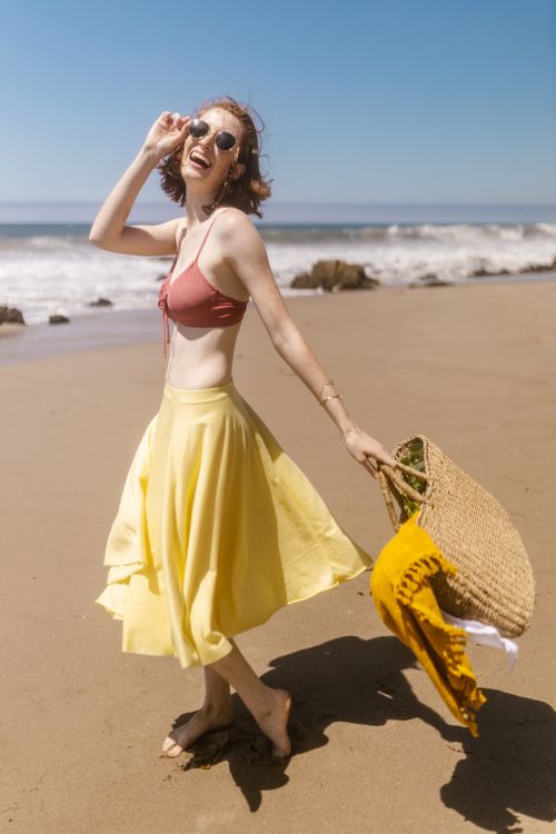 Free Happy woman in a bikini and skirt enjoying a sunny beach day, holding a straw bag and laughing. Stock Photo