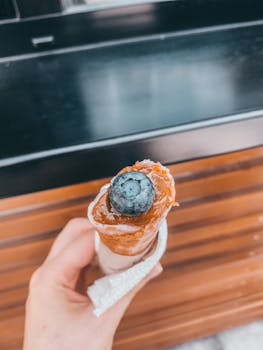 Close-up of a hand holding an ice cream cone topped with a blueberry, showcasing a delicious dessert moment.