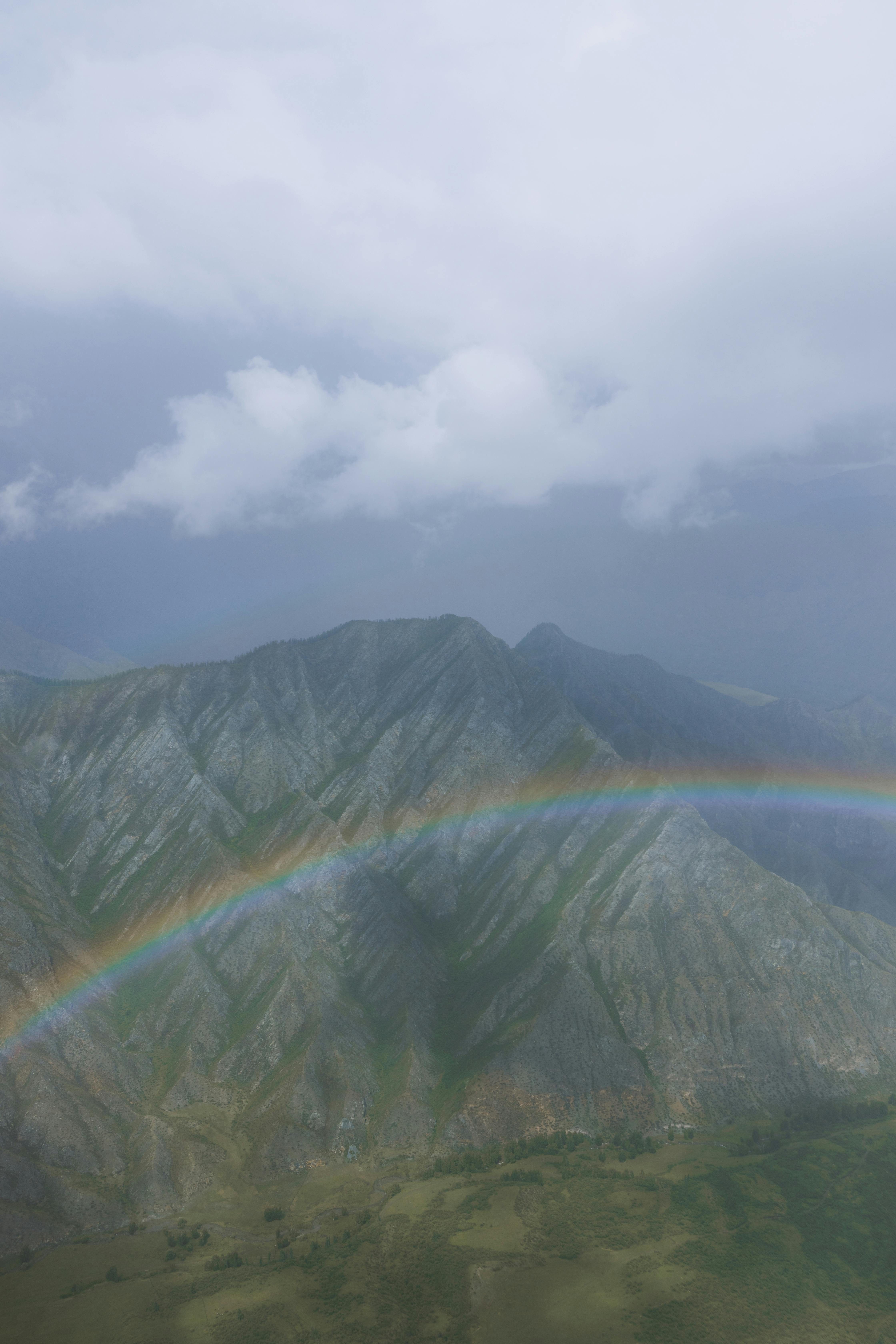 A breathtaking aerial view of a green mountain range with a vibrant rainbow under a cloudy sky.