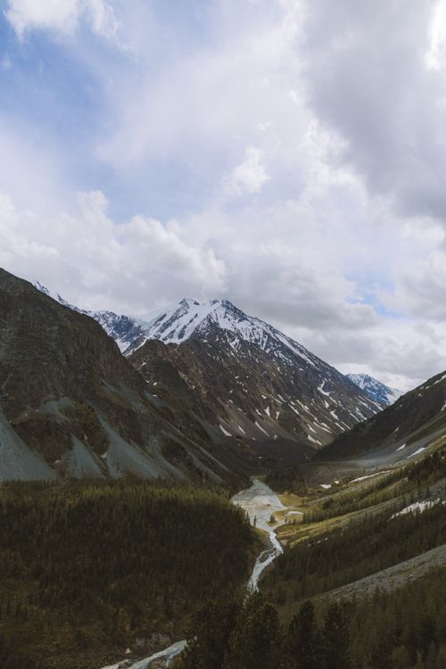 Free A stunning aerial view of a tranquil valley with snow-capped mountains and lush forests under a cloudy sky. Stock Photo