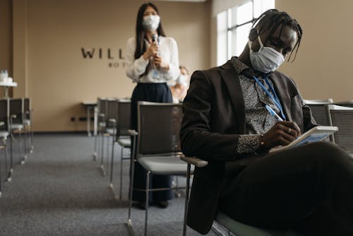 A Man Wearing Blazer and Face Mask Writing on a Spiral Notebook