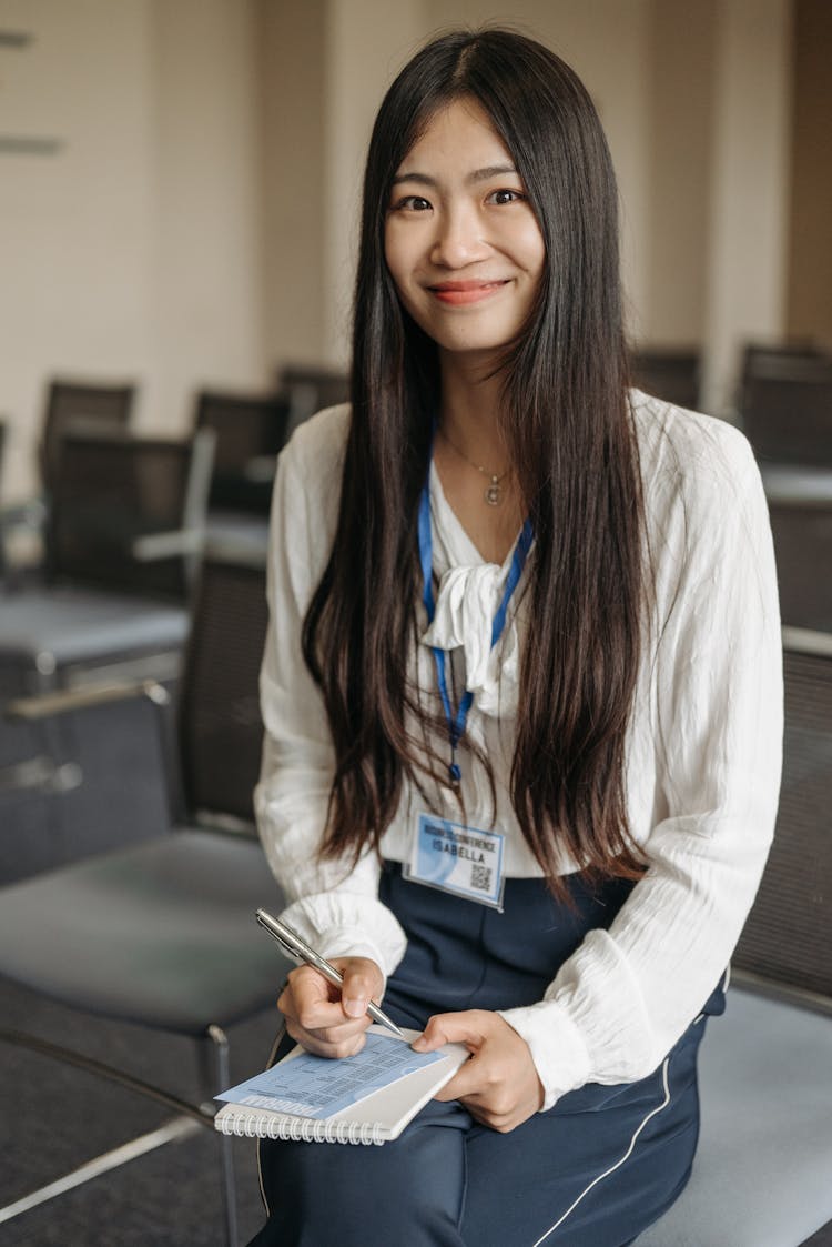 Woman Wearing A White Long Sleeves Top And Badge Holding A Pen And Notebook