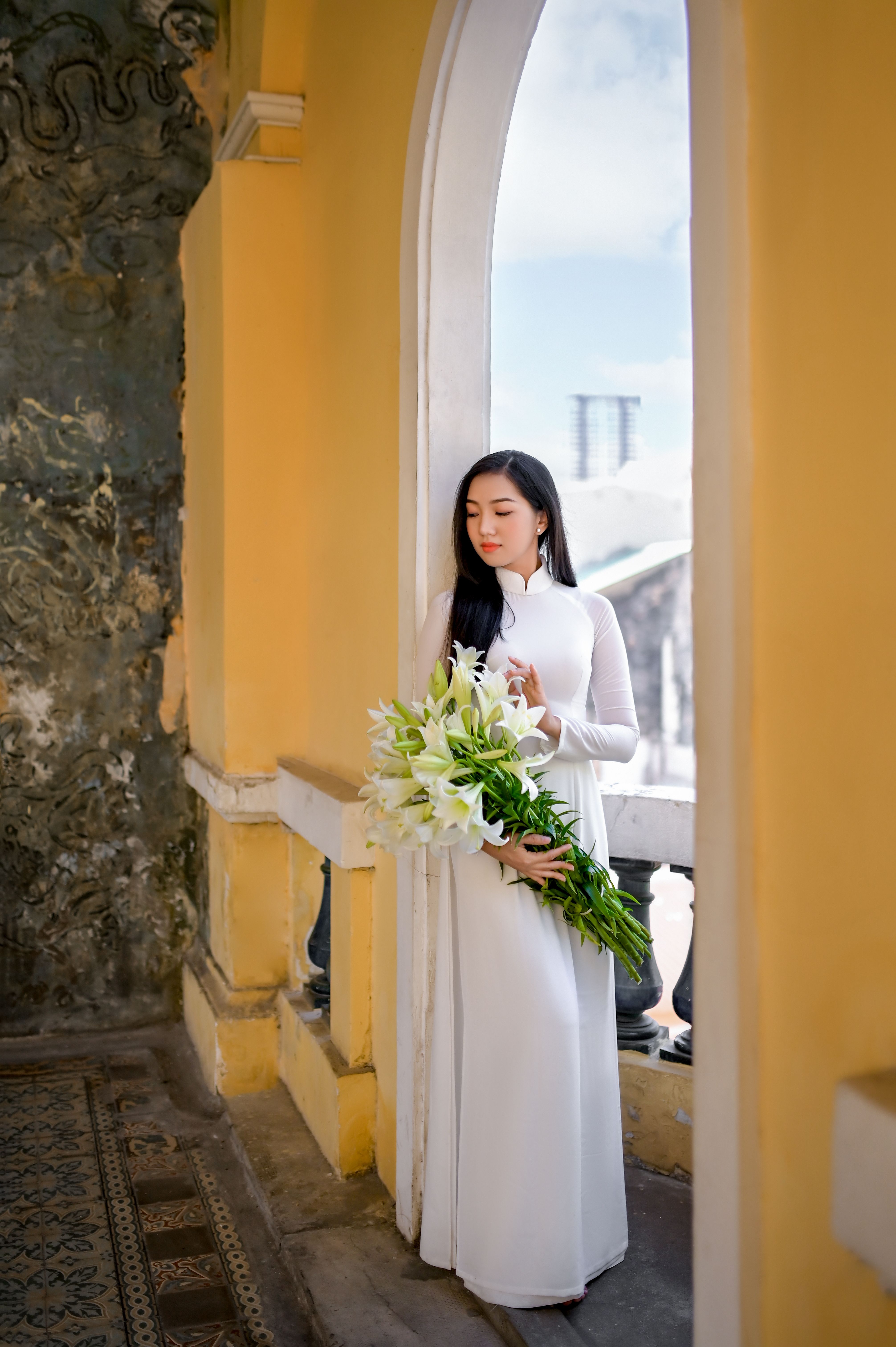 A woman in a traditional Vietnamese gown holding white lilies by a window.