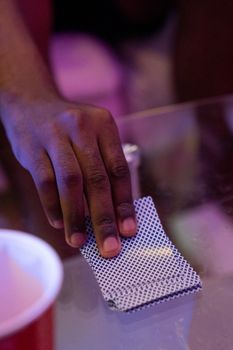 A close-up of a hand holding a deck of playing cards on a glass table with pink lighting.
