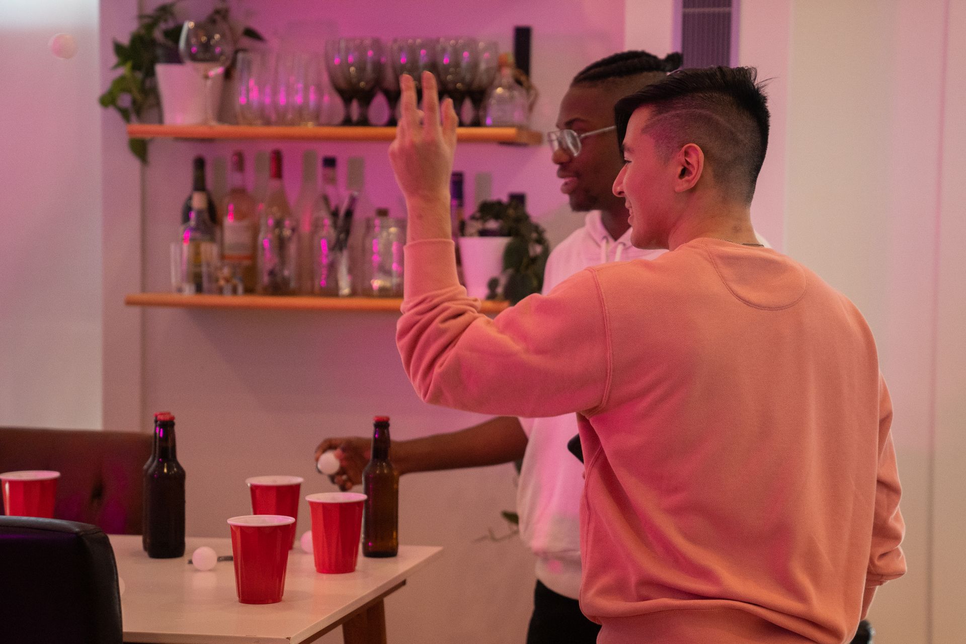 Two friends enjoying a game of beer pong with red cups and bottles indoors.