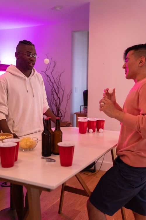 Free Two friends enjoying a fun game of beer pong at an indoor gathering with drinks and laughter. Stock Photo