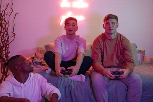 Three young adults enjoy video gaming on a bed in a warmly lit room.