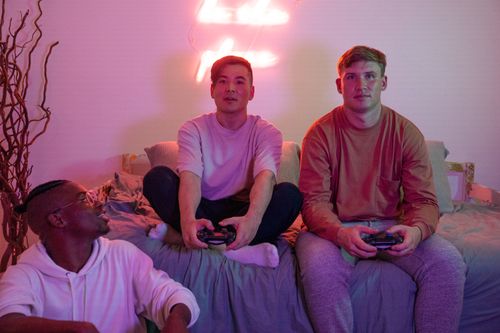 Free Three young adults enjoy video gaming on a bed in a warmly lit room. Stock Photo