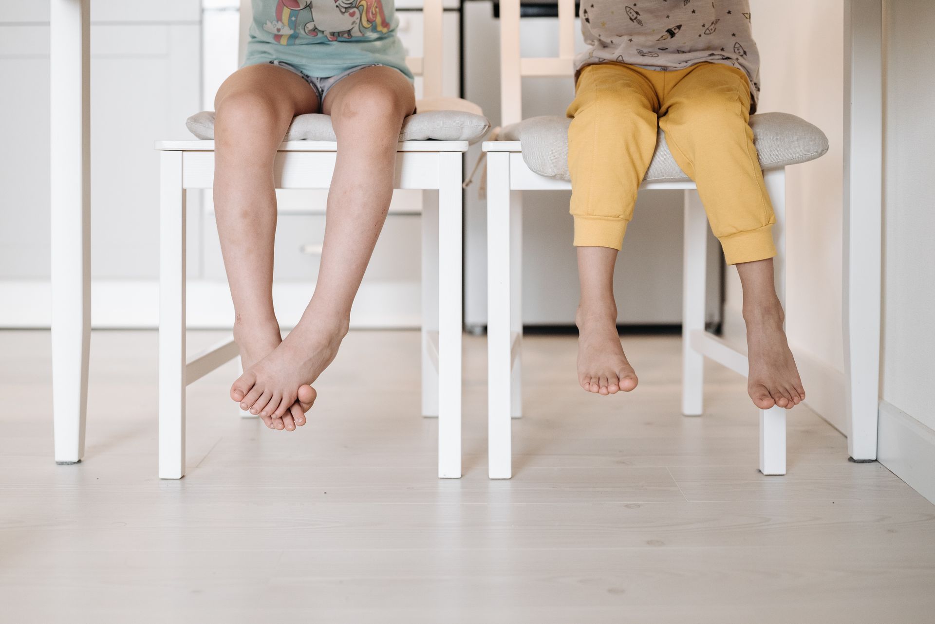 Two children sitting on chairs indoors, feet dangling playfully.