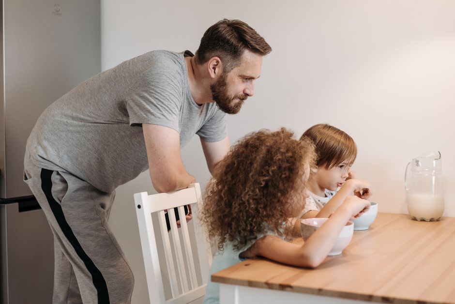 Father checking a shared family calendar on a smartphone while kids play in the background, illustrating coordinated co-parenting and new routines for a multi-child family.. Photographed by Pavel Danilyuk