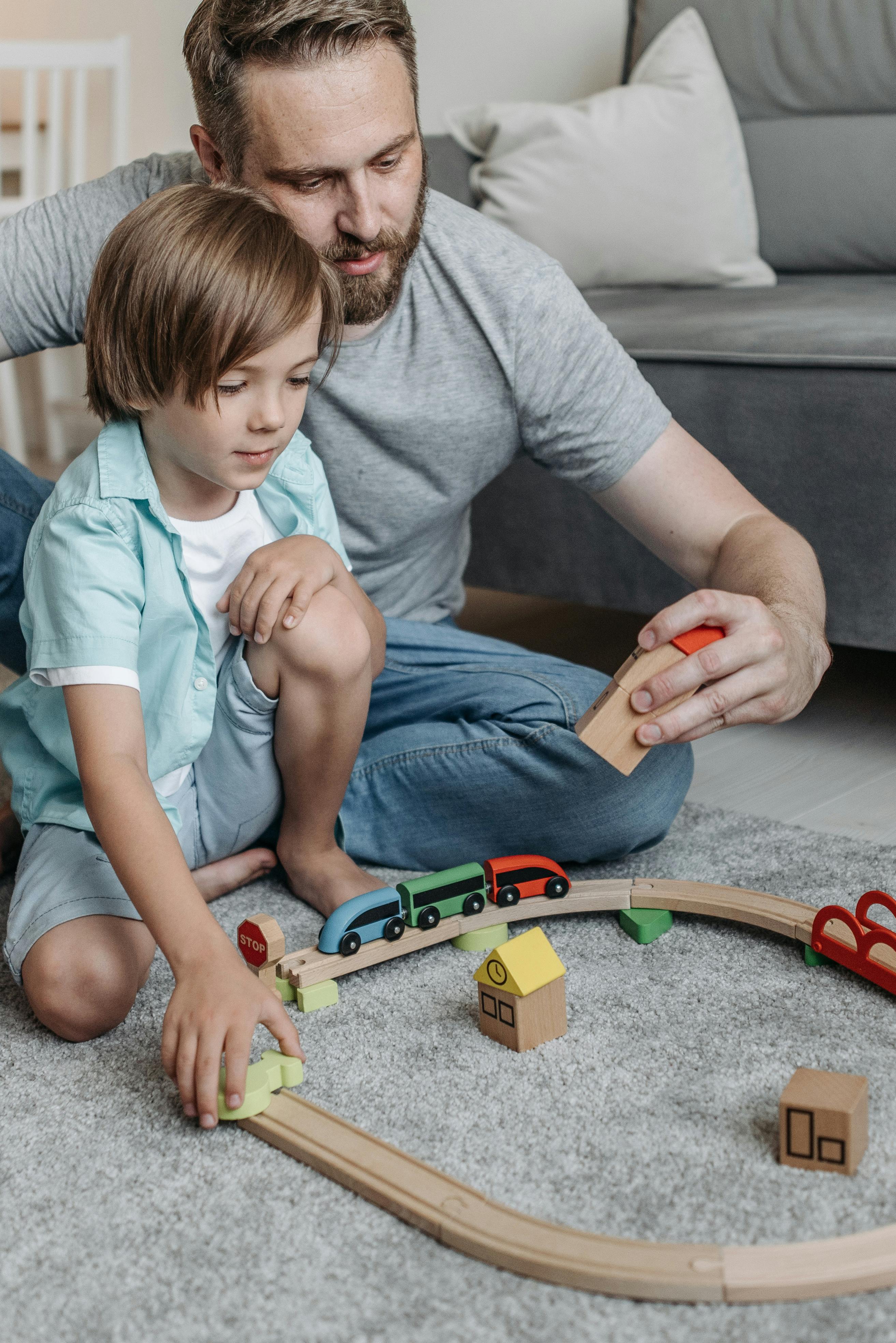 Father and son bonding indoors, playing with a wooden toy train set on the floor.