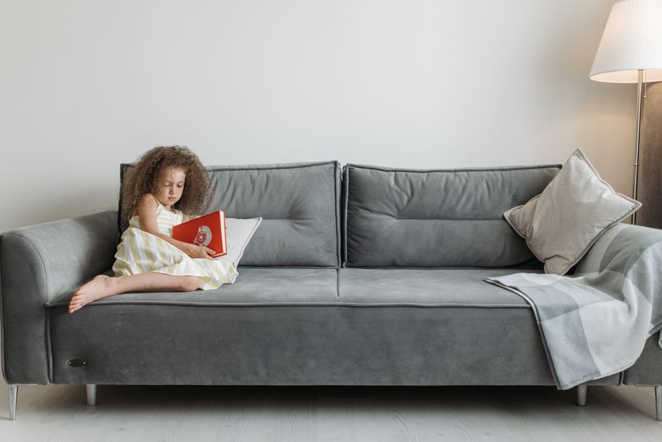 Young girl with curly hair reads a red book on a comfortable grey sofa indoors.