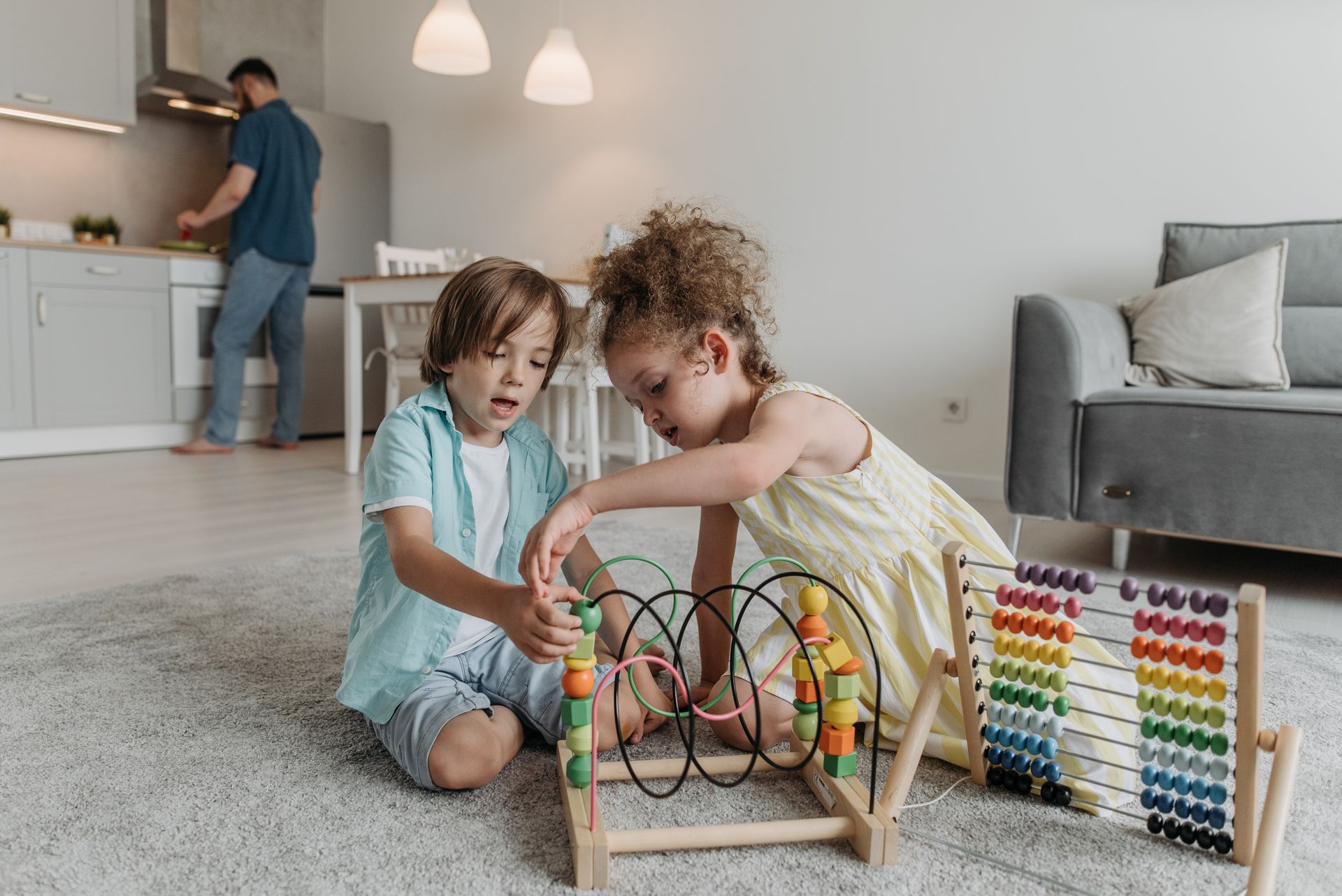 Two children playing with educational toys in a cozy living room while an adult in the kitchen.