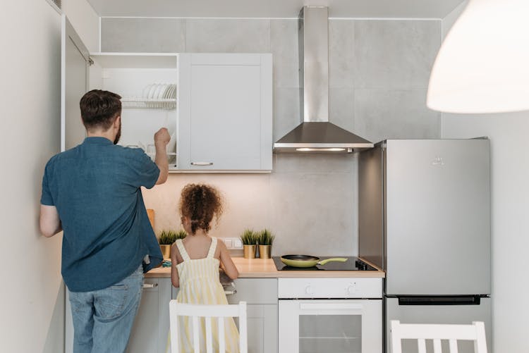 A Man Standing Beside A Little Girl Getting Plates From The Wooden Cabinet Of The Kitchen 