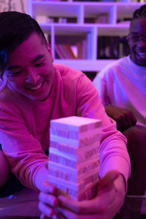 Free Two friends enjoying a game of Jenga indoors with colorful ambient lighting. Stock Photo