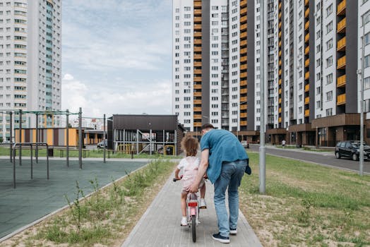A dad helps his daughter learn to ride a bike on a pathway among tall residential buildings.