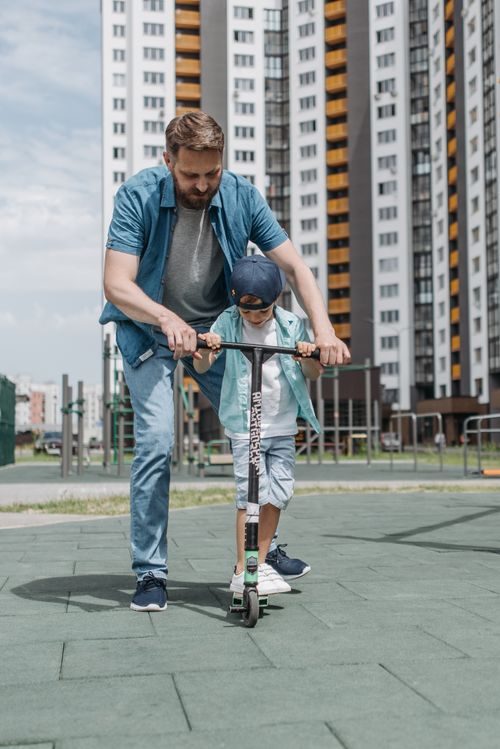 Free A father guides his son on a scooter in an urban park with modern buildings in the background. Stock Photo