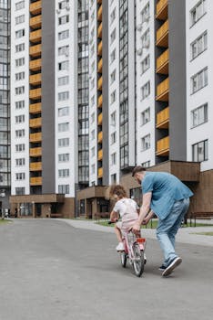 A father guides his daughter on a bike ride near towering apartment buildings.