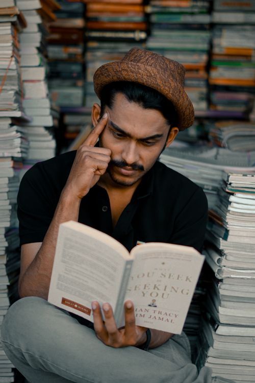 Free A thoughtful man in a hat reads a book in a bookstore surrounded by stacks of books. Stock Photo