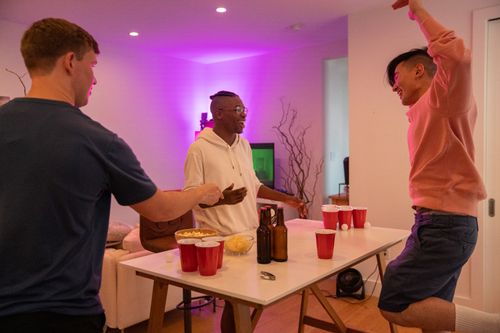 Free Three friends having fun playing beer pong indoors with drinks and snacks. Stock Photo
