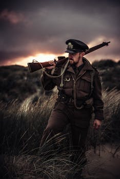 日落时分，一名身着制服的士兵持步枪走过绿草茵茵的沙丘