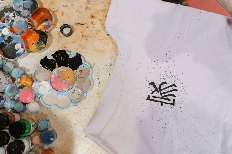 Top view of a t-shirt with artistic design surrounded by colorful paint supplies.