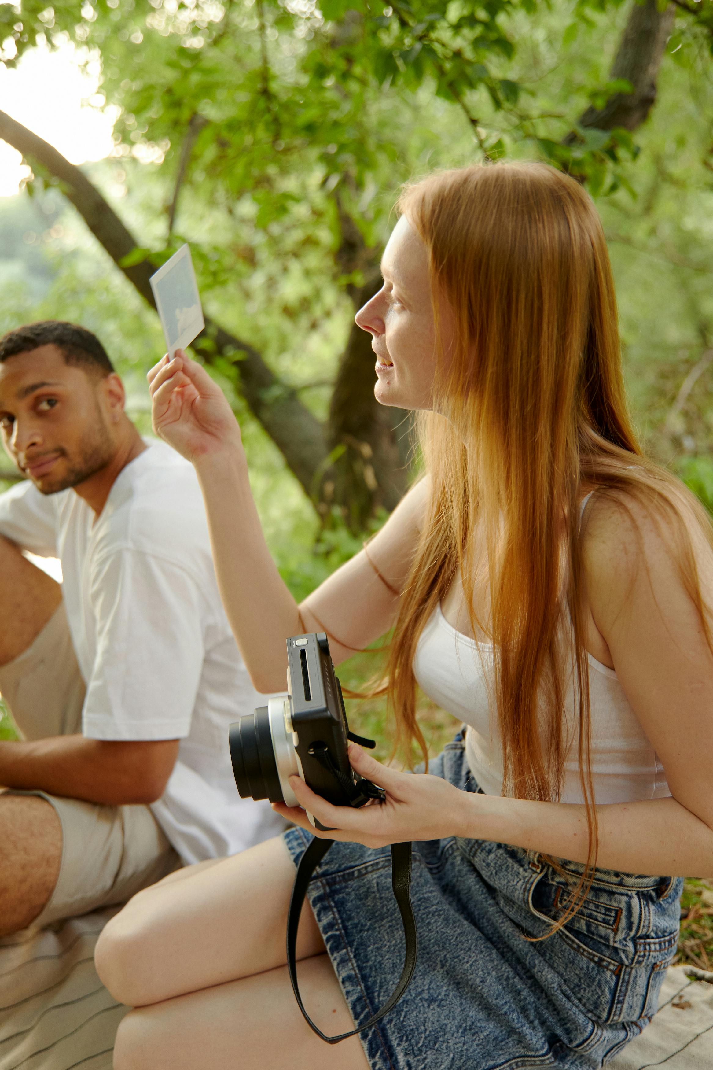 Two friends sitting outdoors, capturing moments with a Polaroid camera in a serene forest setting.