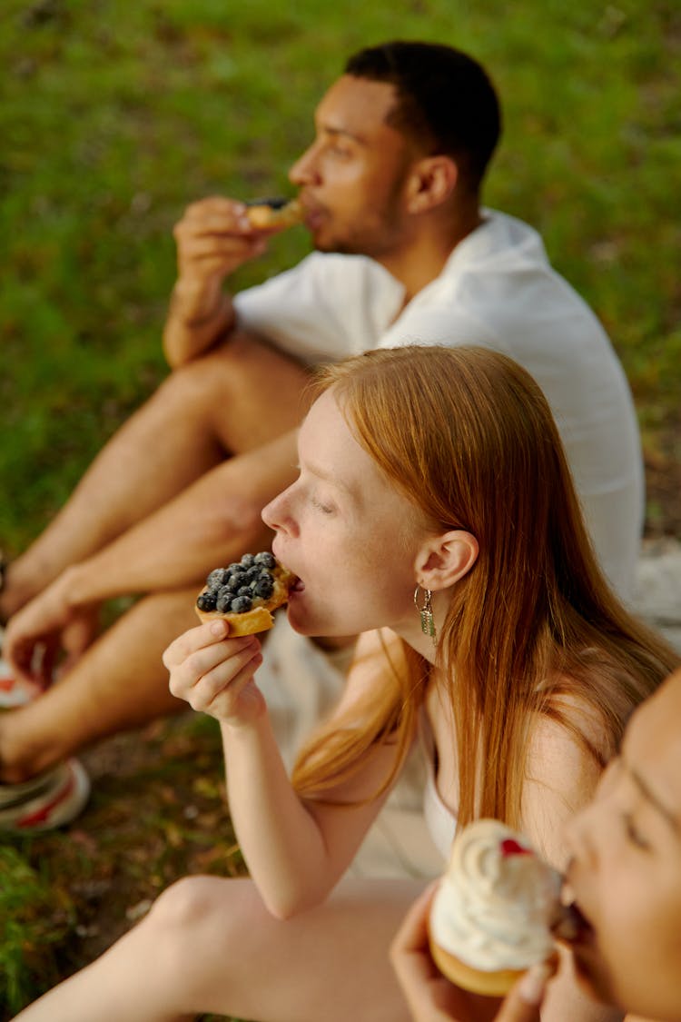 A Man And Women Sitting On The Grass While Eating Pastries