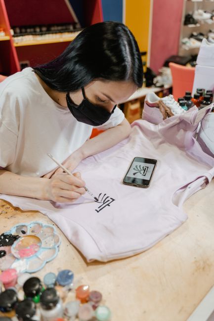 Artist meticulously hand-painting a design on a t-shirt, using a smartphone for reference.