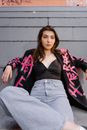 Woman in Red and Black Floral Cardigan and Gray Denim Jeans Sitting on Gray Concrete Wall