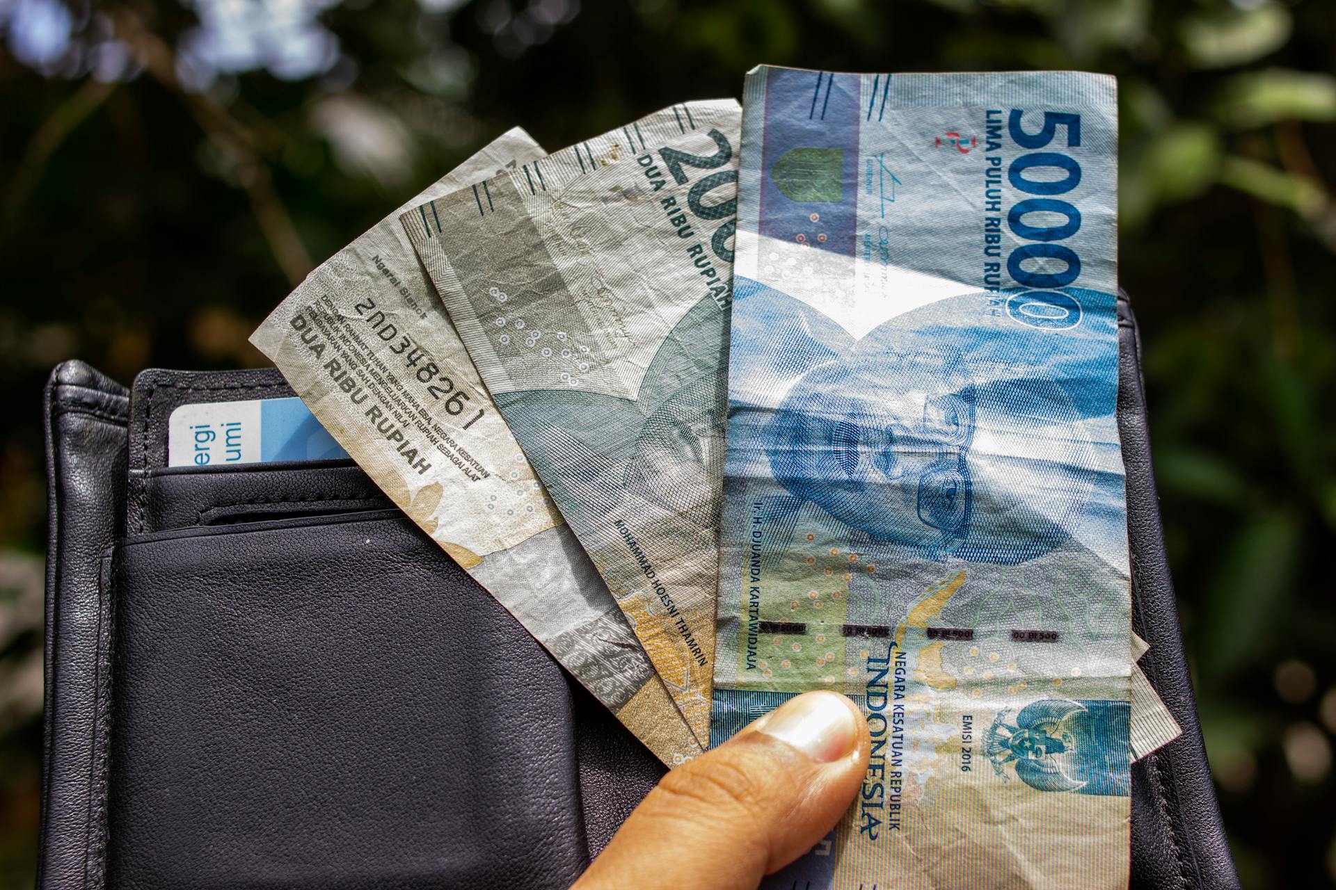 Close-up of a hand holding Indonesian Rupiah banknotes outdoors with a wallet.