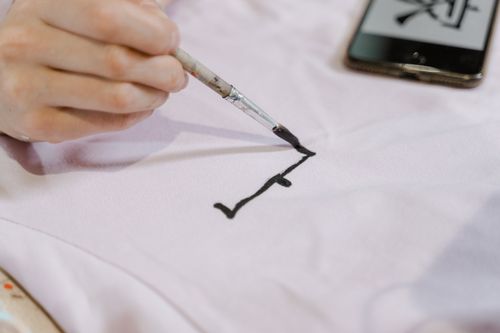 Free Detailed close-up of a hand using a brush to paint on white fabric, near a smartphone. Stock Photo