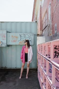 Fashionable woman in sunglasses stands in an urban alleyway with graffiti and pastel colors, showcasing modern street style.