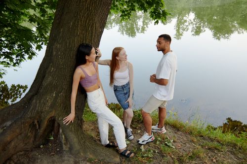 Free Three friends relaxing and chatting by a riverside tree on a summer day. Stock Photo