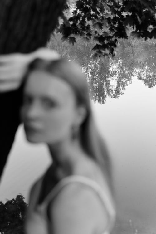 Grayscale Photography of a Woman Leaning on a Tree Trunk Near a Lake