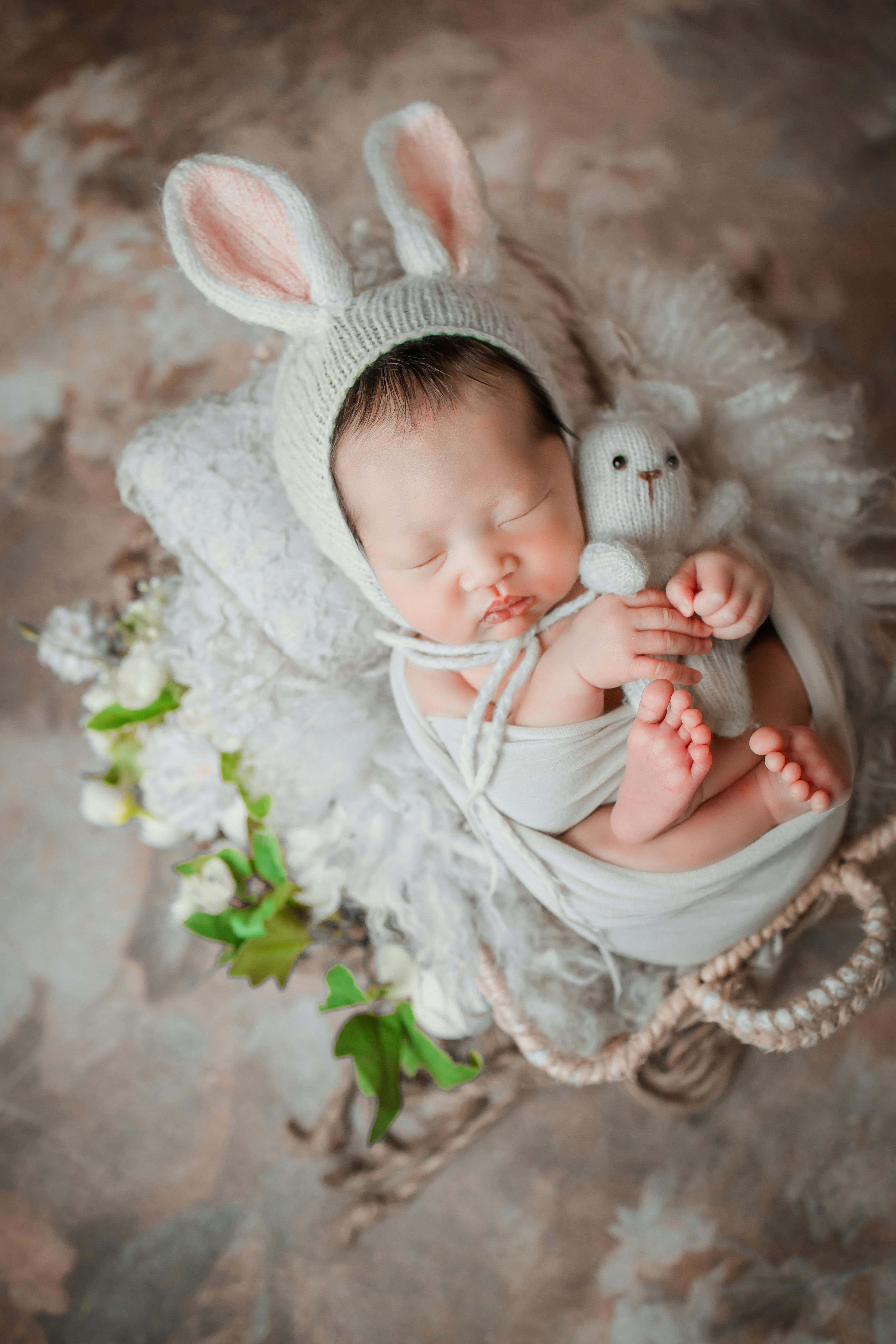 An adorable newborn baby sleeping peacefully with bunny ears hat and plush toy, surrounded by soft textures and flowers.