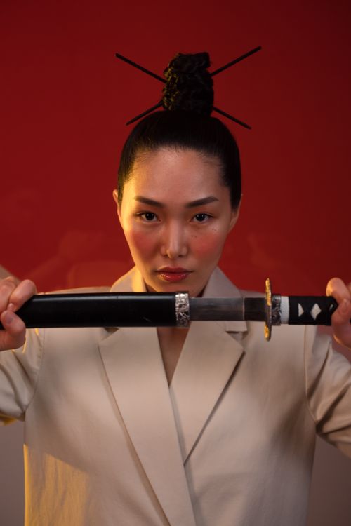 Free Focused Asian woman wearing a white blazer, holding a katana, showcasing a serious expression indoors. Stock Photo