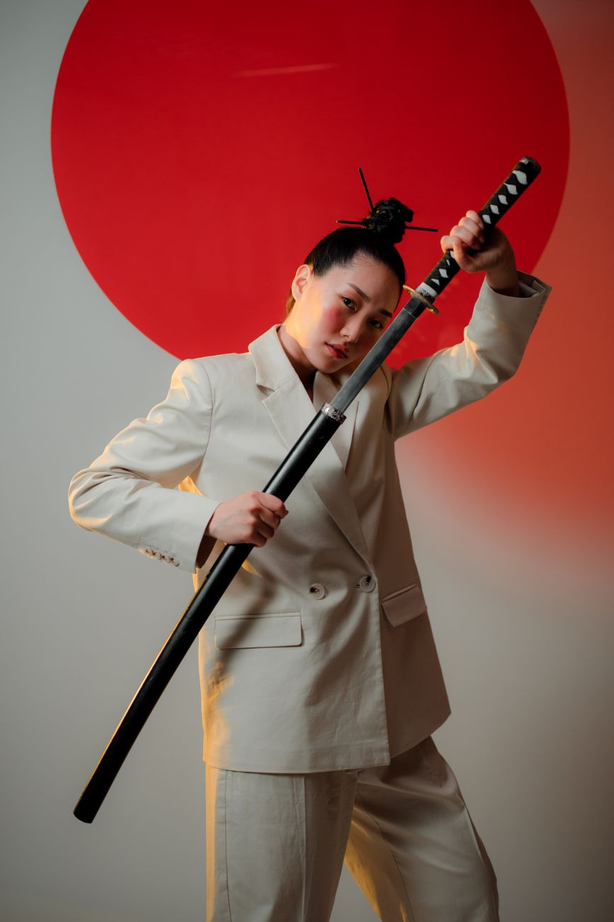 Striking portrait of an elegant woman in a blazer, confidently posing with a katana sword against a bold red circle back