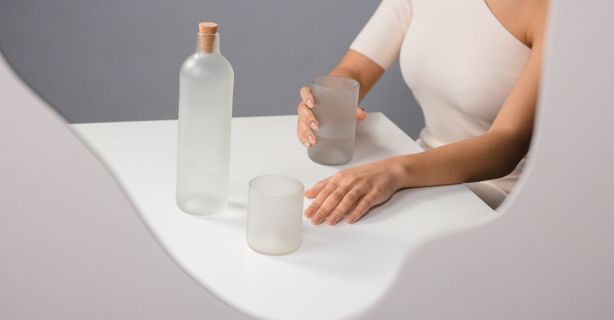 Composição minimalista elegante com uma garrafa de vidro fosco e copos para beber, destacando a mão de uma mulher.
