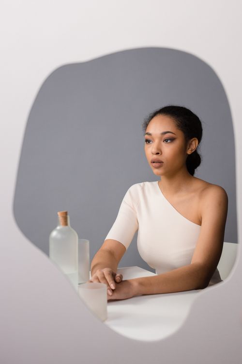 Free A woman in an off-shoulder dress reflects calmly in a uniquely shaped mirror with minimalist decor. Stock Photo