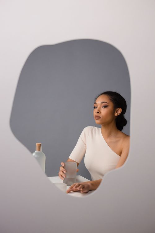 Free Sophisticated woman in white holding a glass, seen through a reflective mirror cutout. Stock Photo