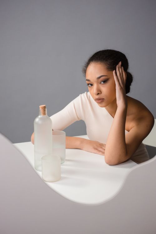 Free A sophisticated studio portrait of a woman with glass bottles on a minimalist white table. Stock Photo
