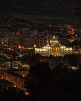 Nighttime view of a brightly lit university building in Tirane, Albania with surrounding cityscape.