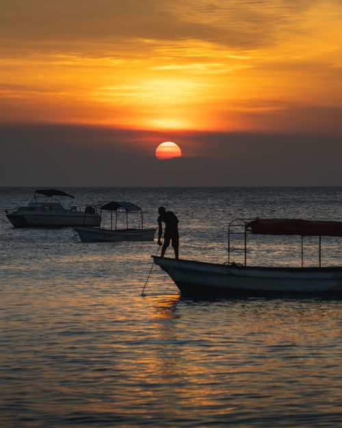 Free Serene beach sunset with silhouetted fisherman and boats on calm sea. Perfect for travel and nature themes. Stock Photo