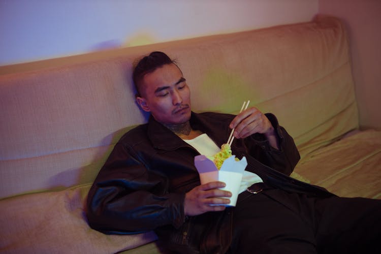 A Man Sitting On A Couch Eating Using Chopsticks