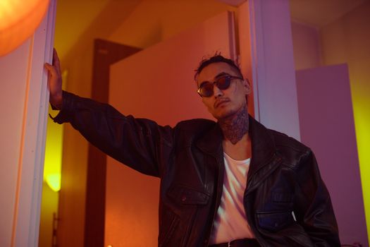 Confident man wearing sunglasses and a leather jacket stands in a dimly lit room.