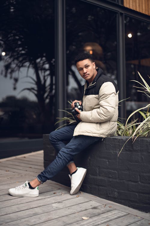 A Man in a Puffer Jacket Holding His Camera while Sitting on a Concrete Planter