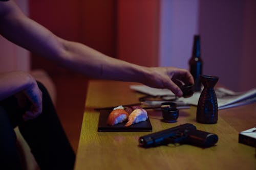 Close-up of Man Reaching for an Item on a Table with Sushi and a Gun 